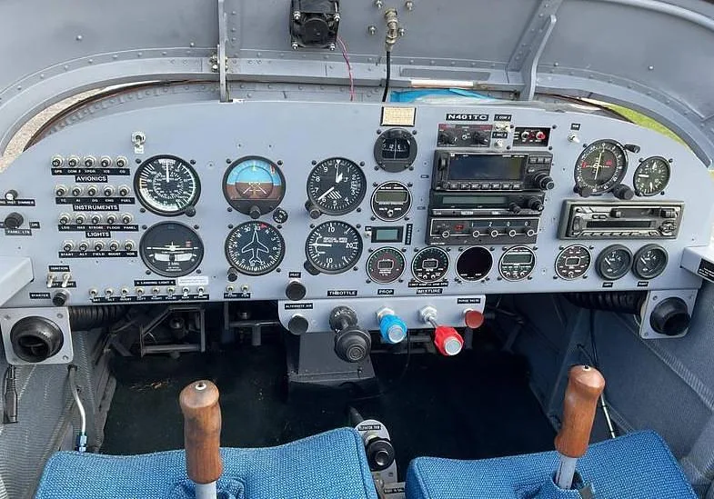 The cockpit of an aircraft, showing the instrument panel with various gauges, radios, and controls, and two blue seats.
