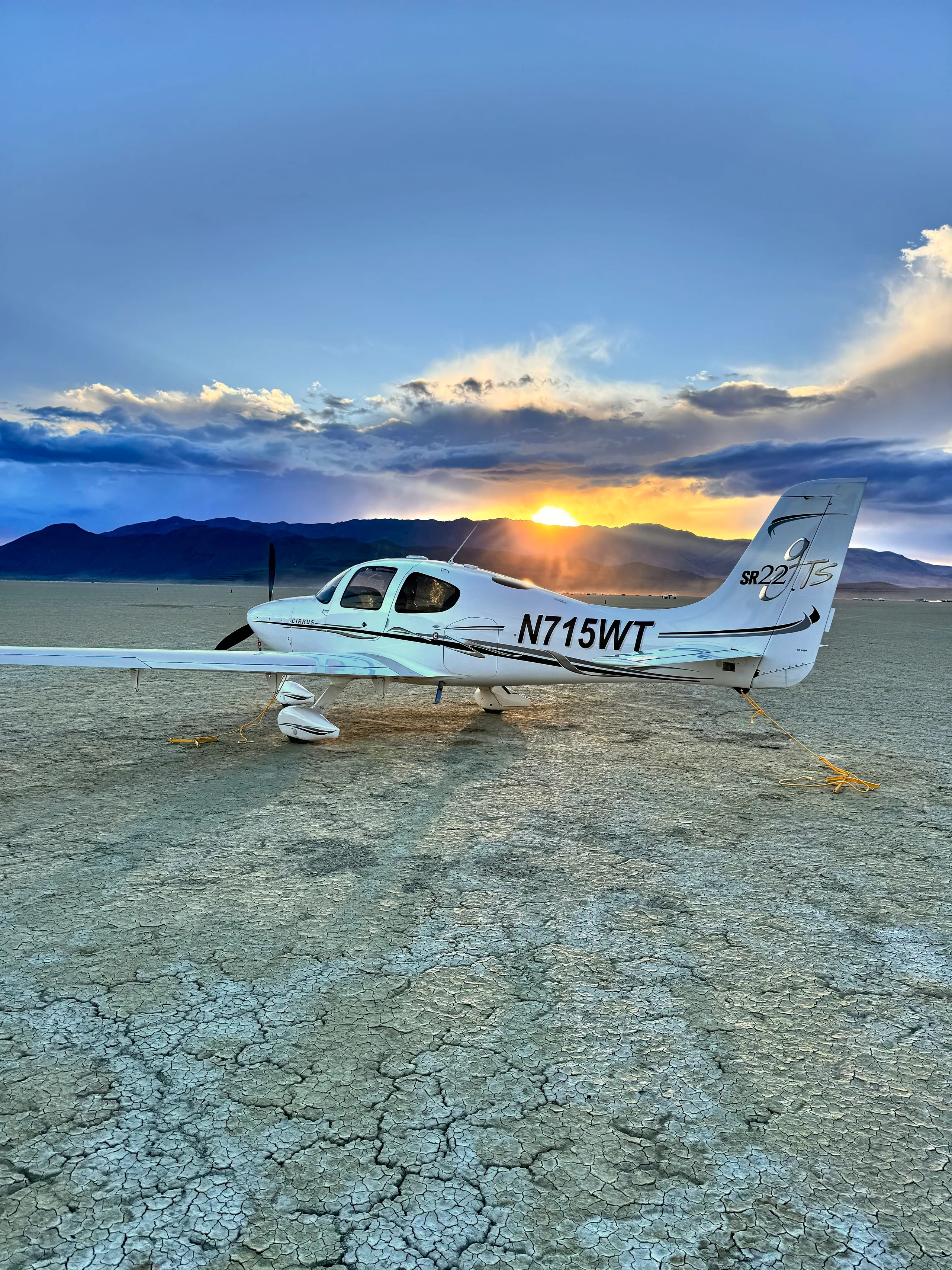 A white Cirrus SR22T aircraft (N715WT) parked on a cracked dry lake bed at sunset, with sun rays beaming from behind.