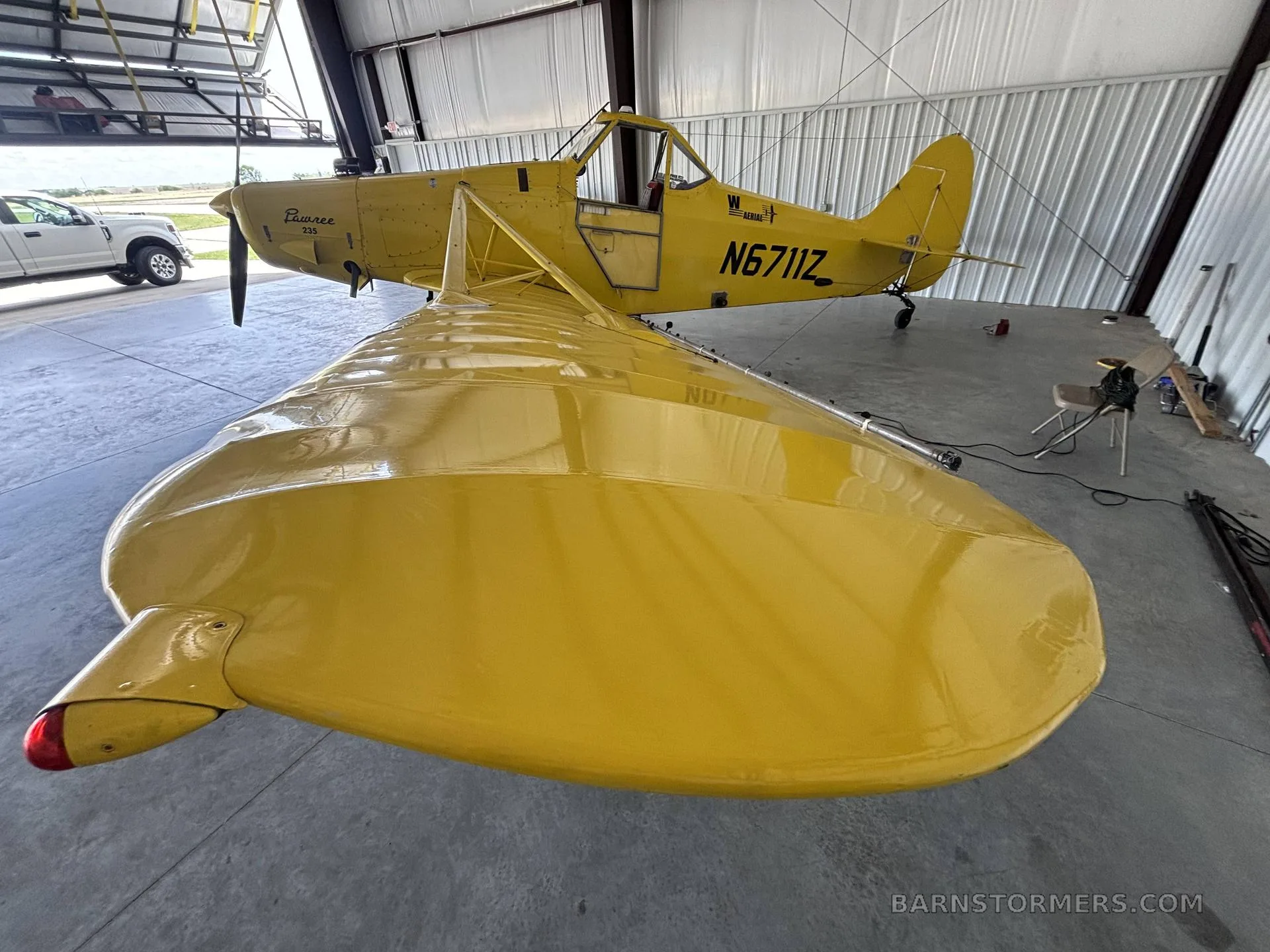 A yellow Piper PA-25 Pawnee aircraft with registration N6711Z, viewed from the rear-right in a hangar.
