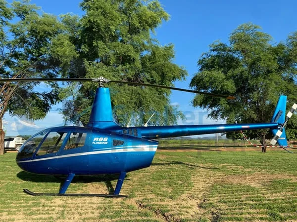 A blue Robinson R66 Turbine helicopter with white stripes is parked on a grassy field with trees in the background.