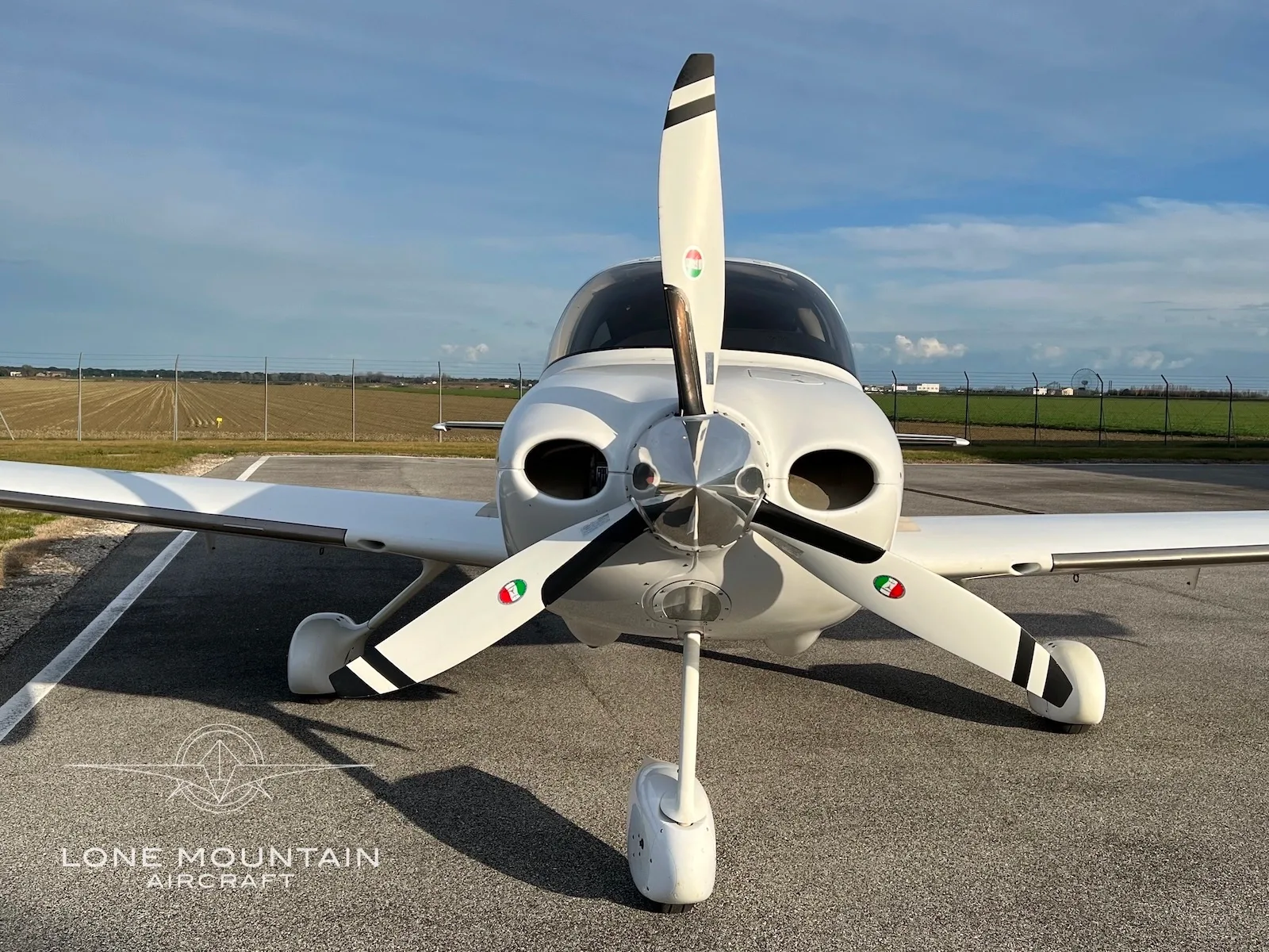 A head-on view of a white Cirrus SR22 aircraft with Italian flag colors on its propeller blades, parked on an airfield.