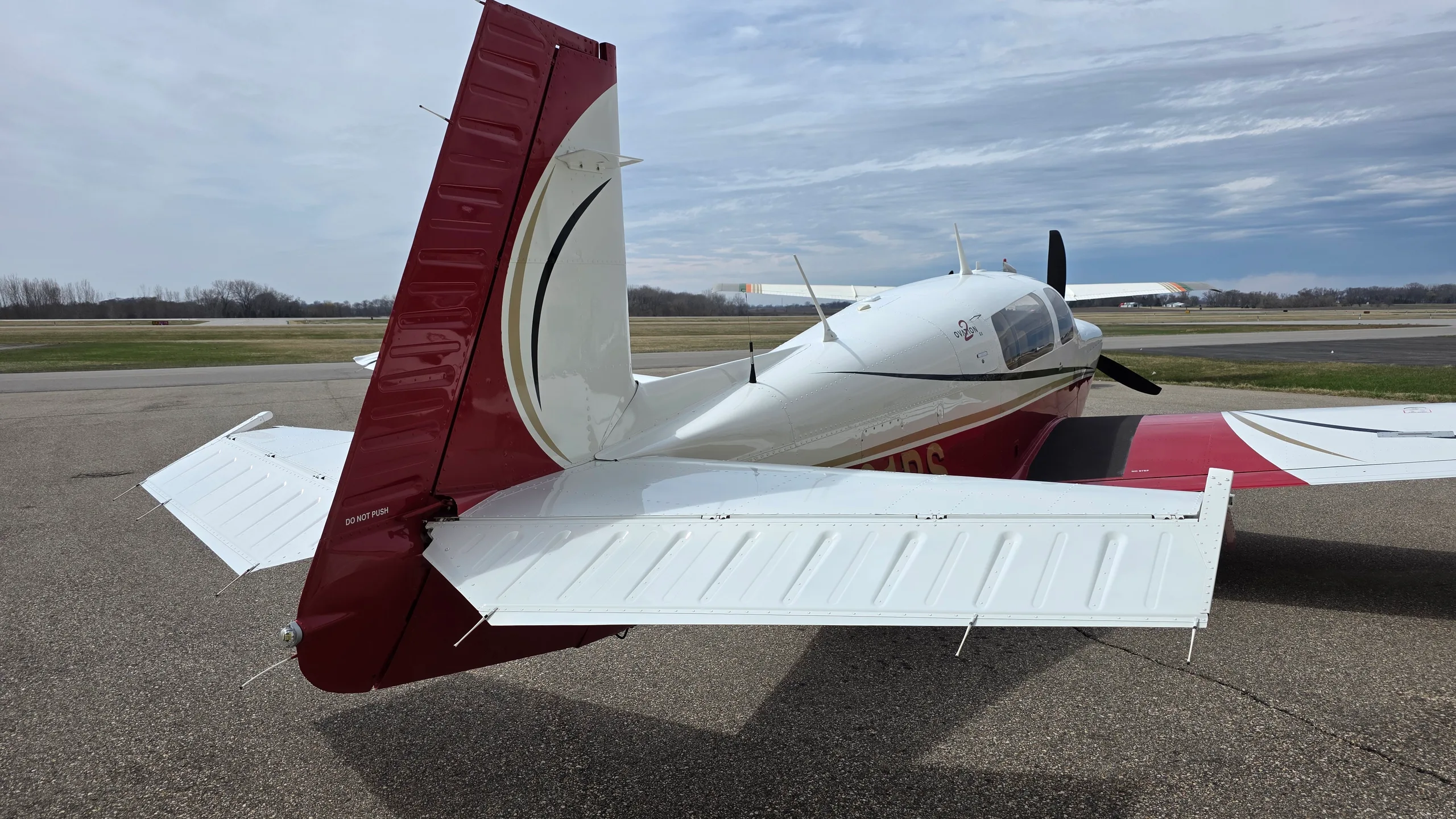 A rear view of a red and white Mooney aircraft with its tail and horizontal stabilizers visible on an asphalt apron.
