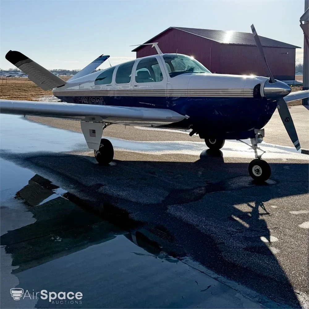 Front-left view of a blue and white Beechcraft Bonanza on a wet tarmac with a hangar in the background.