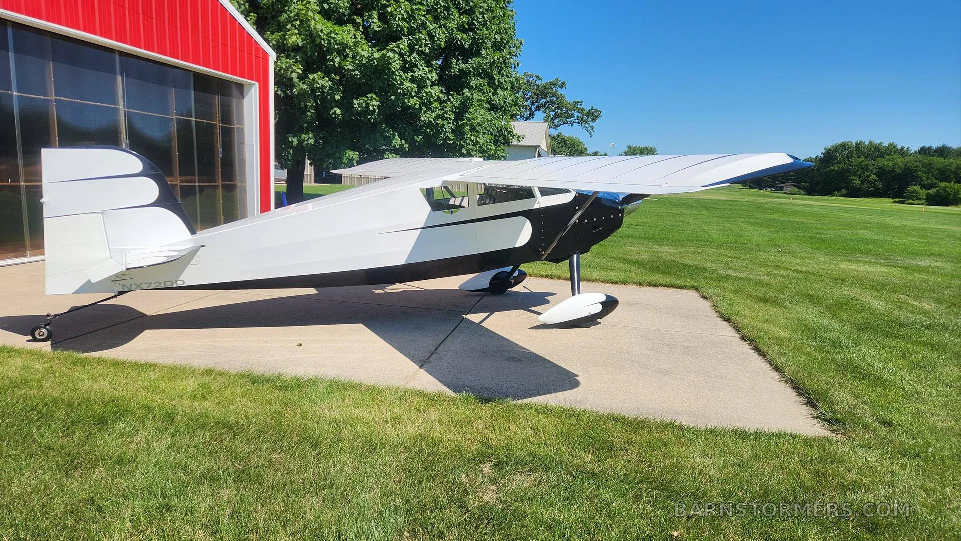 A black and white single-engine aircraft with tail number NXZ2DD is parked on concrete next to a red hangar.