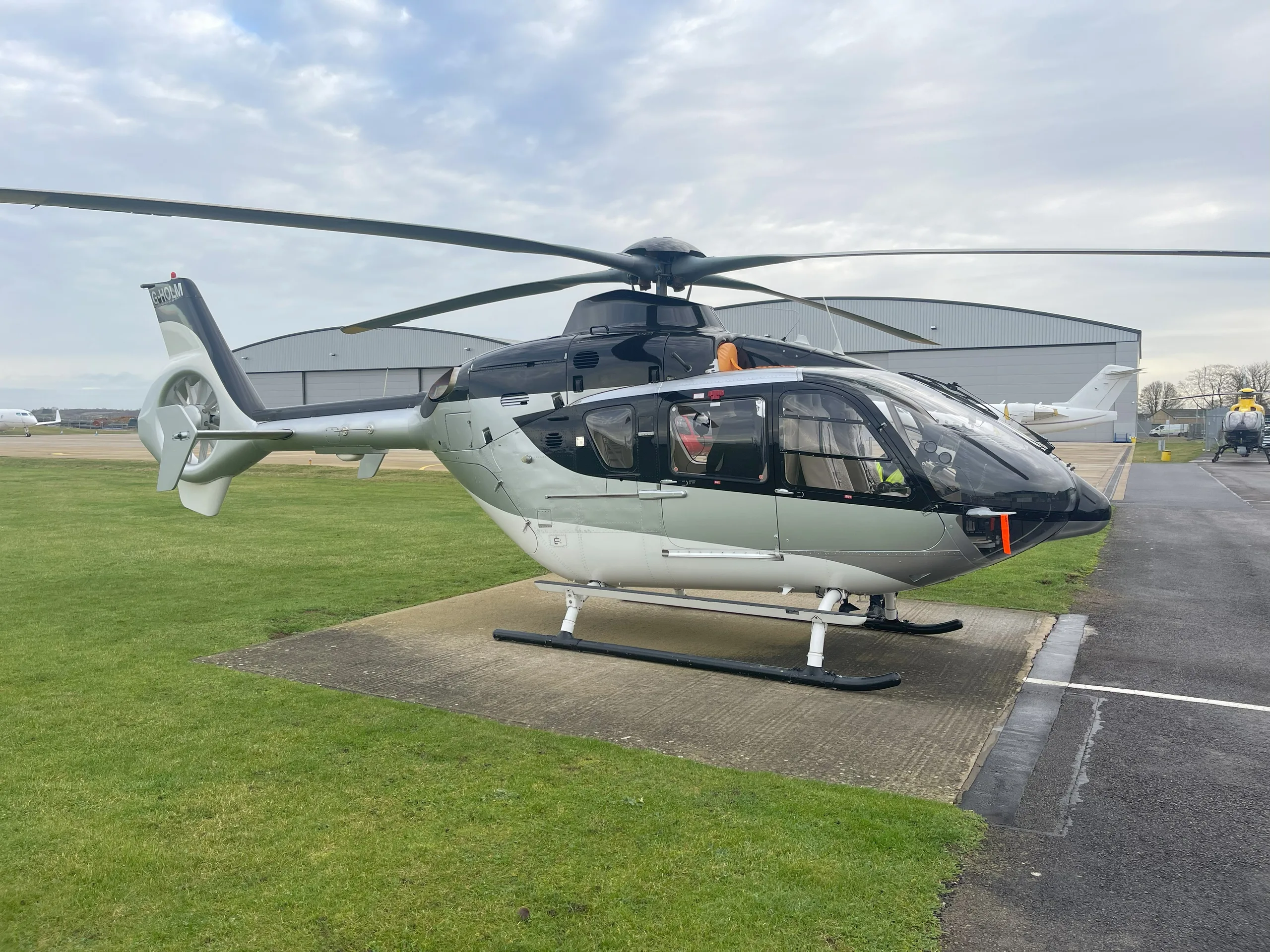 A modern black, grey, and white helicopter (registration G-HOLM) is parked on a concrete pad at an airfield under a cloudy sky.