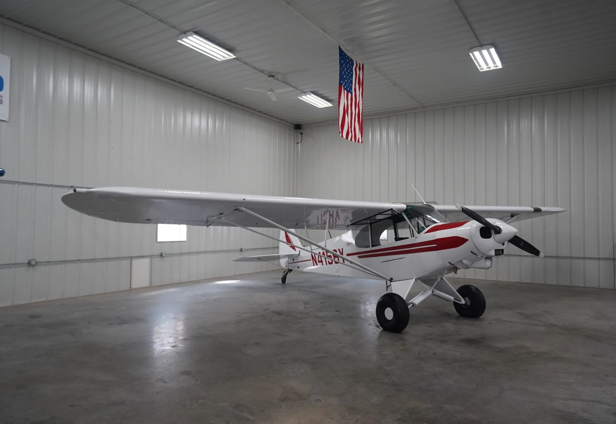 A white Piper Cub aircraft, registration N4158Y, with red stripes is parked in a hangar with an American flag hanging overhead.