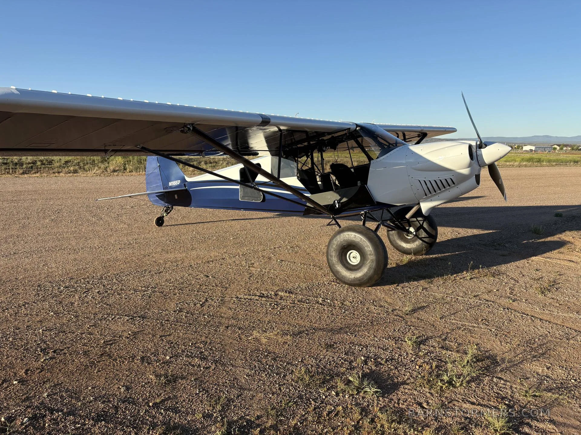 A side view of a white and blue CubCrafters aircraft with large tundra tires parked on a dirt field.