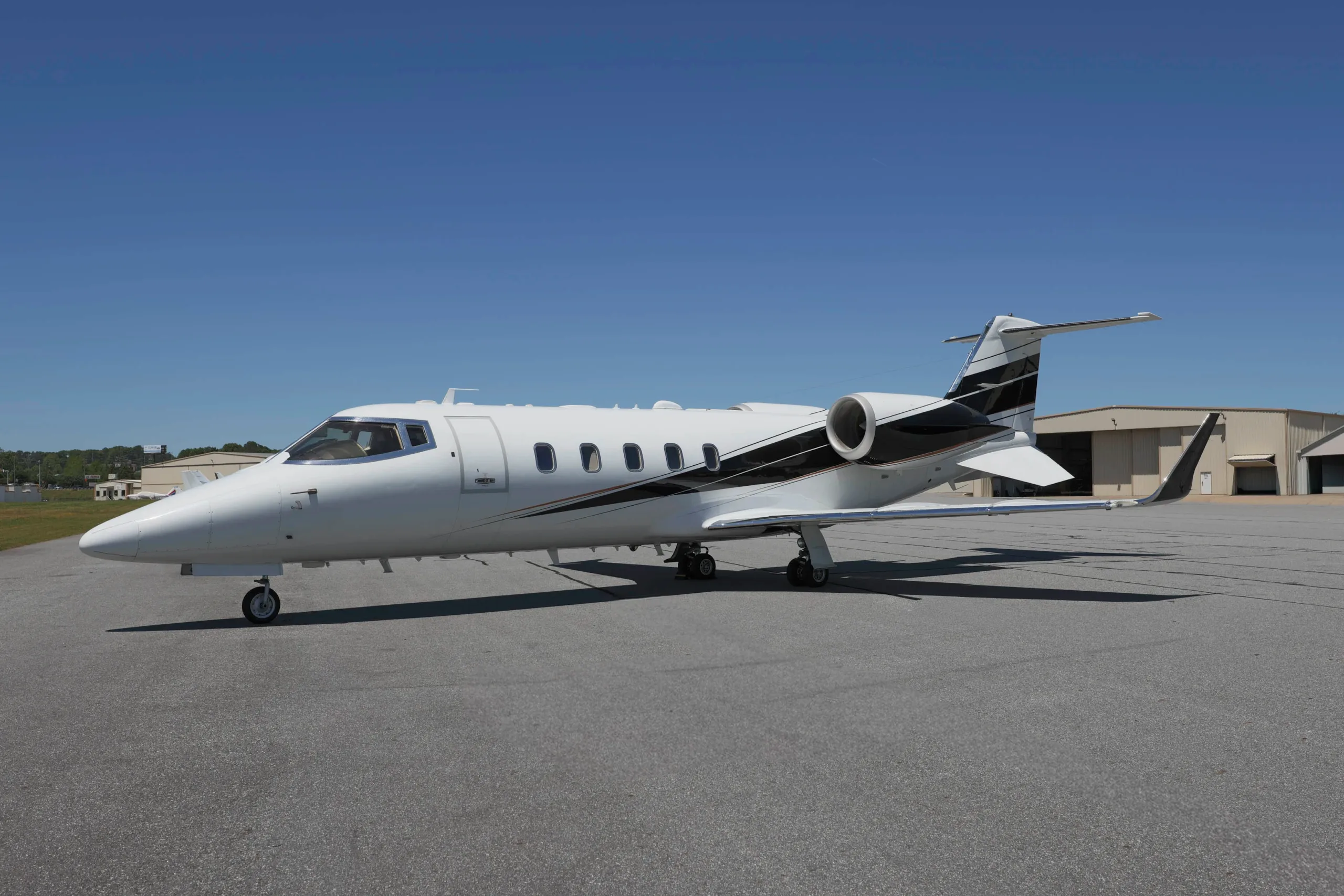 A white business jet with black and gold stripes, viewed from the side on an airport tarmac with hangars in the distance.