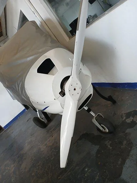 Front view of a white light aircraft with a two-bladed propeller and nose wheel, partially covered by a grey tarp, inside a hangar.