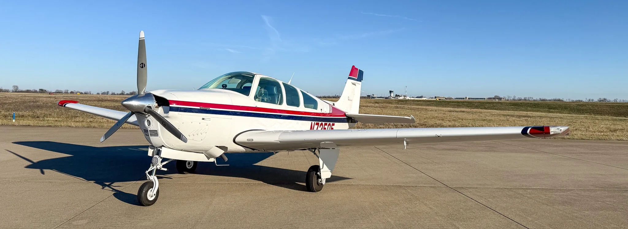 Front-left view of a white Beechcraft Bonanza with red and blue stripes on an airfield under a clear blue sky.