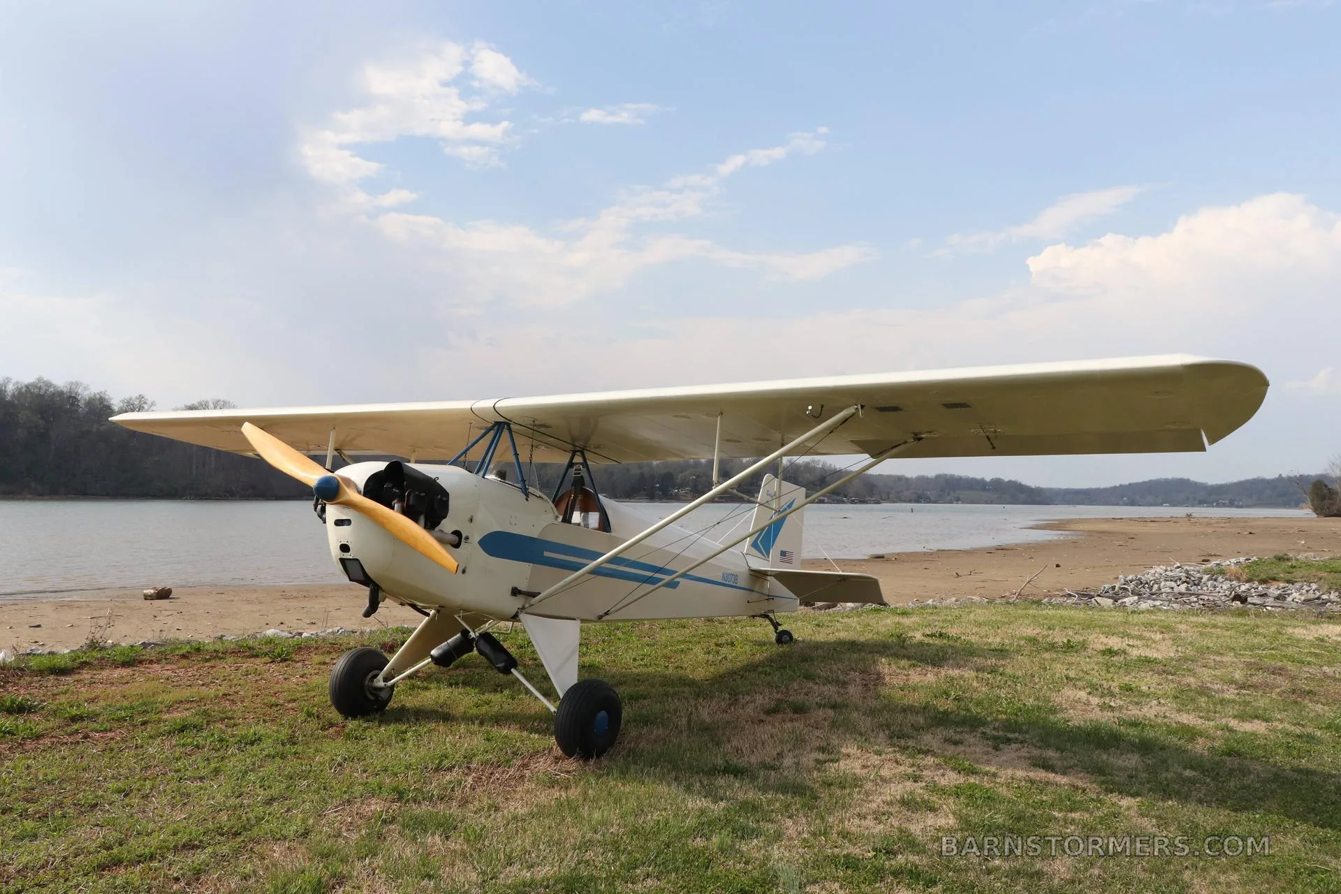 A white and blue single-engine taildragger aircraft, N3073B, with a wooden propeller, parked on grass by a lake.