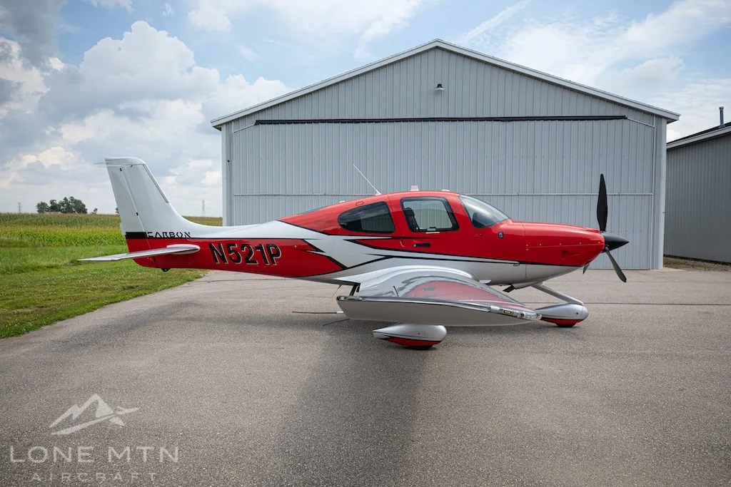 A red and silver Cirrus SR22 aircraft, N521P, parked on an asphalt ramp next to a hangar.