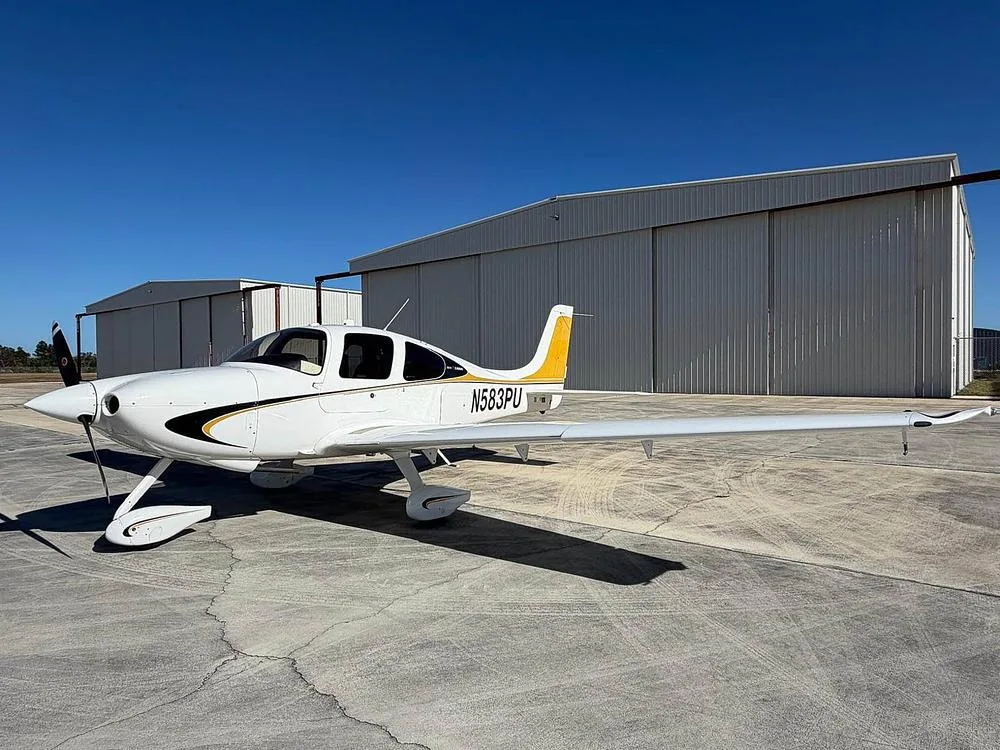 A white Cirrus SR22 aircraft with black and gold stripes and a gold tail, registration N583PU, parked on an airfield with hangars in the background.