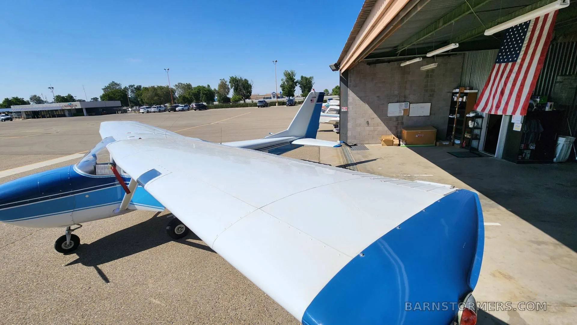 A blue and white Cessna aircraft is parked on an airfield next to a hangar with an American flag, viewed from above the right wing.