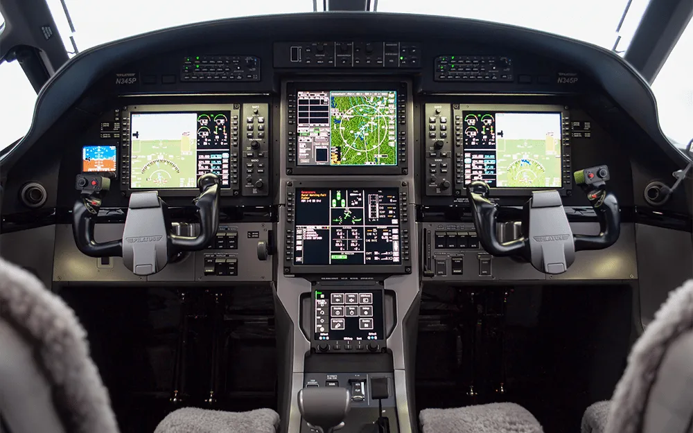 The modern glass cockpit of a Pilatus PC-12 NGX, featuring multiple large display screens and control yokes.