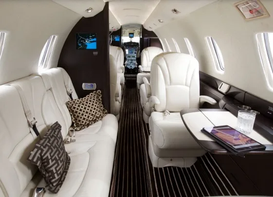 The luxurious interior of a private jet features white leather seats, dark wood accents, and a patterned aisle carpet.