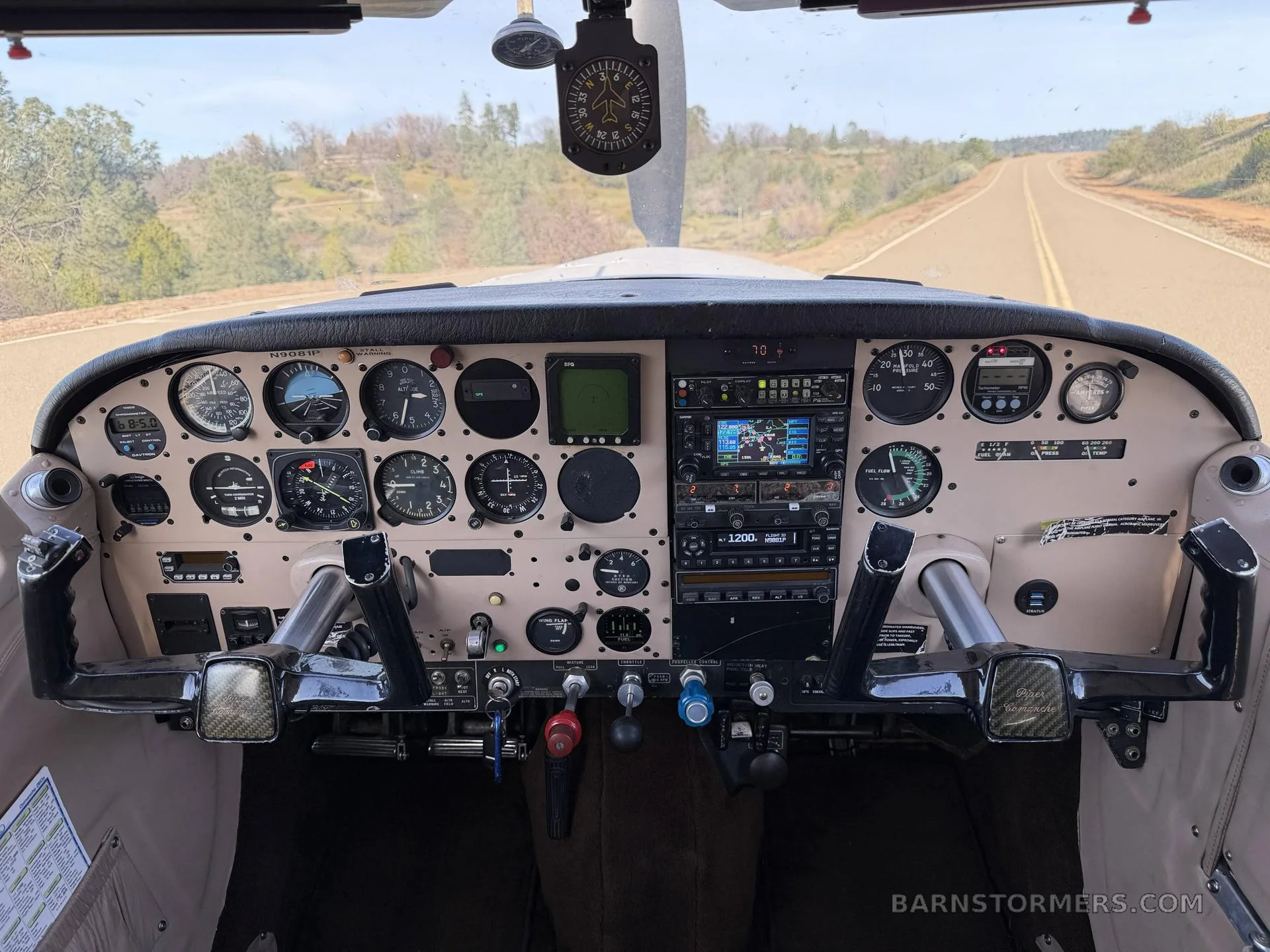 The cockpit of a small aircraft shows the instrument panel, yokes, and controls, with a road visible through the windshield.