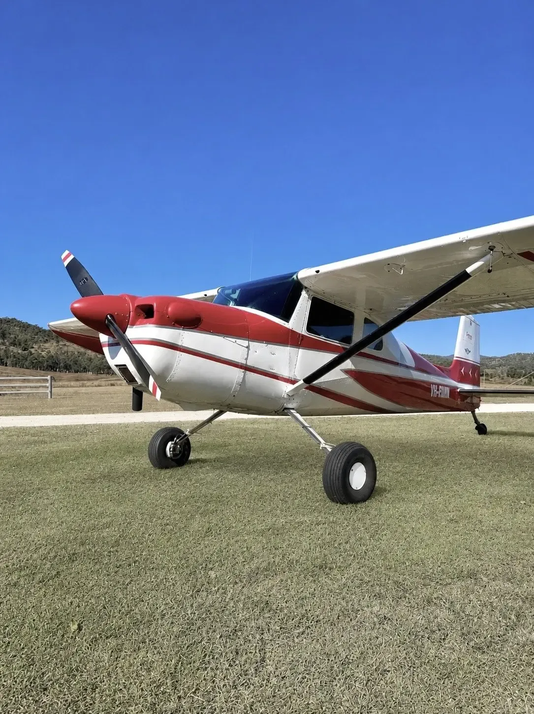 A red and white single-engine propeller aircraft with registration VH-RWM is parked on a grassy field under a clear blue sky.