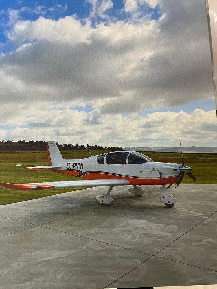 A side view of a white and orange light aircraft with registration ZU-PVW, parked on a concrete pad under a cloudy sky.