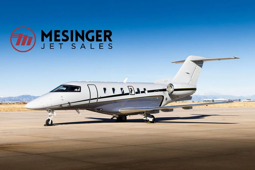 A gray and black business jet, likely a Phenom 300, parked on an airfield with mountains in the background.