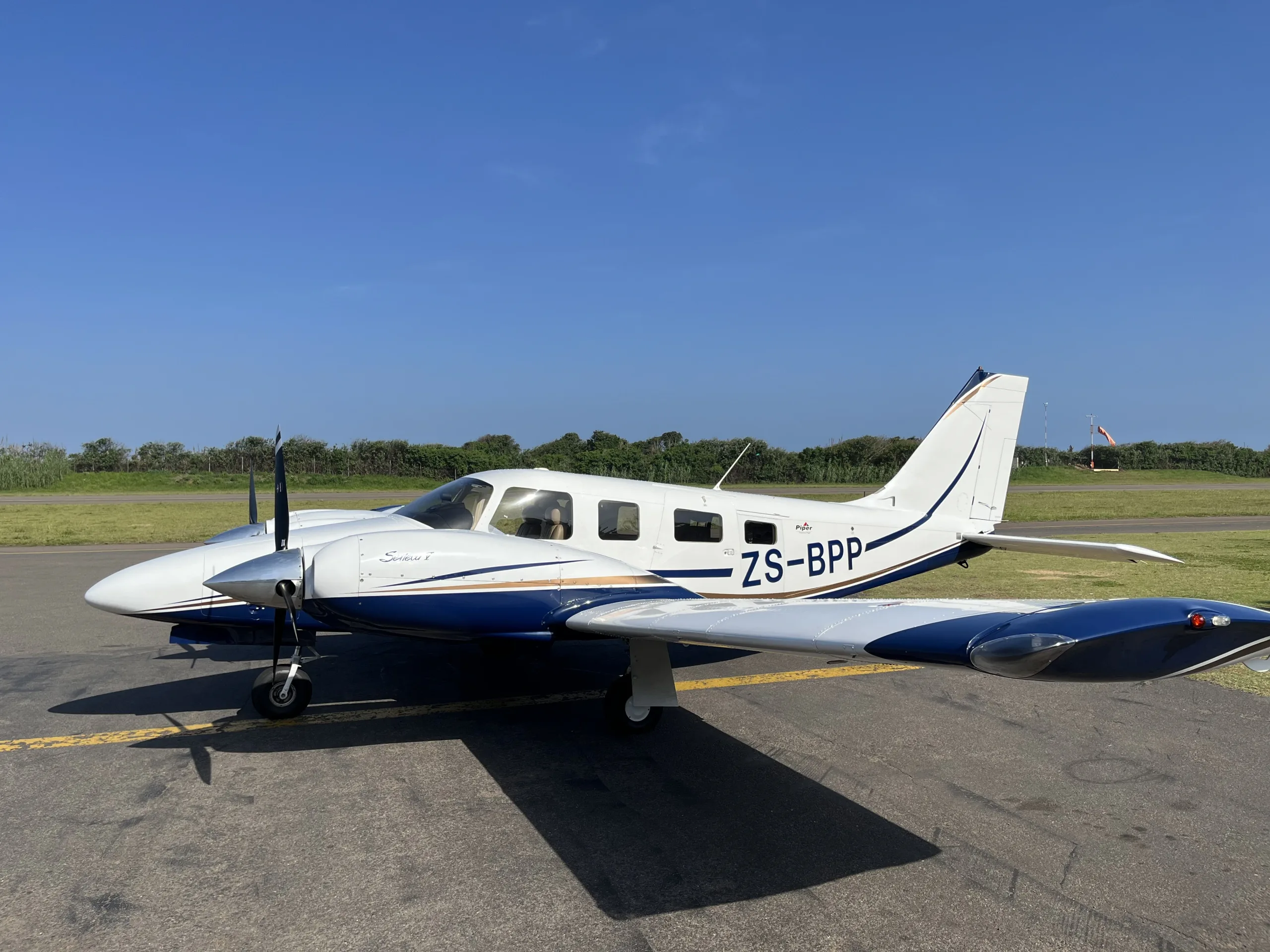 A white and blue Piper Seneca twin-engine aircraft, registered ZS-BPP, is parked on an airfield under a clear blue sky.
