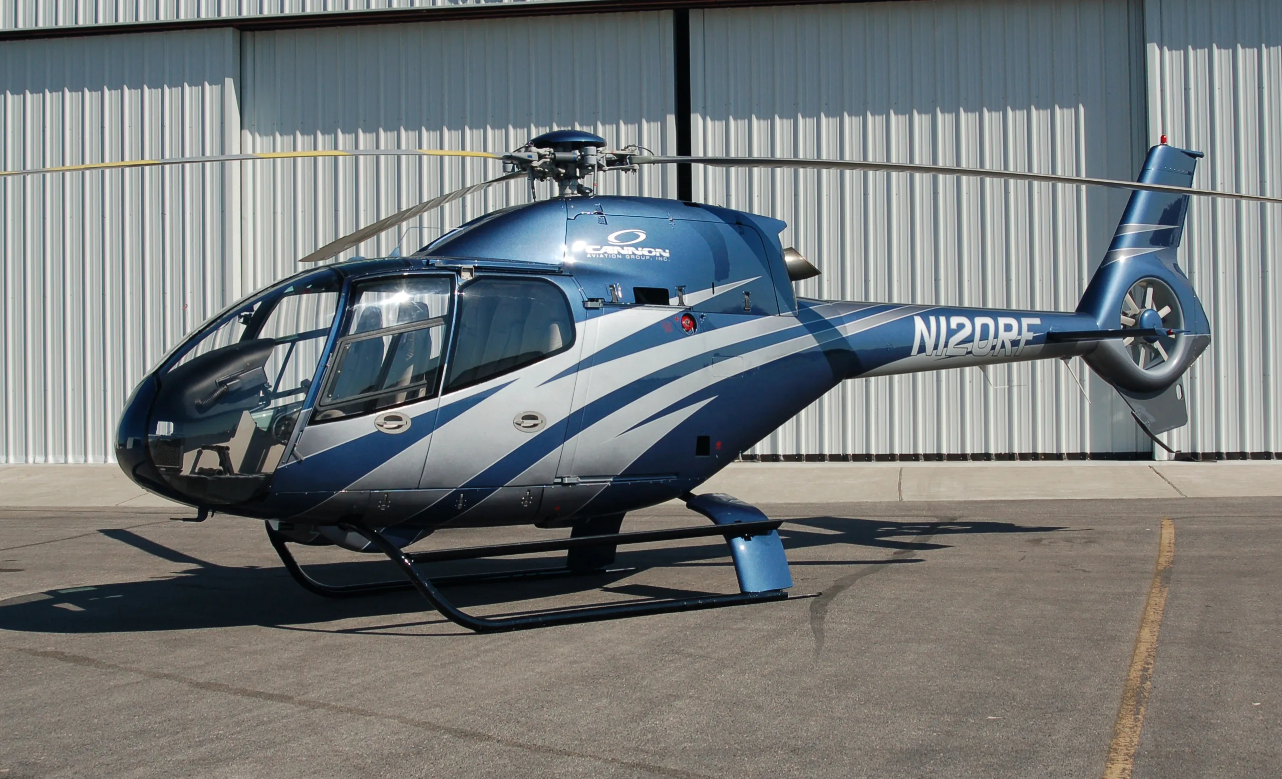 A blue and silver Eurocopter EC120 Colibri helicopter with registration N12DRE parked in front of a hangar.