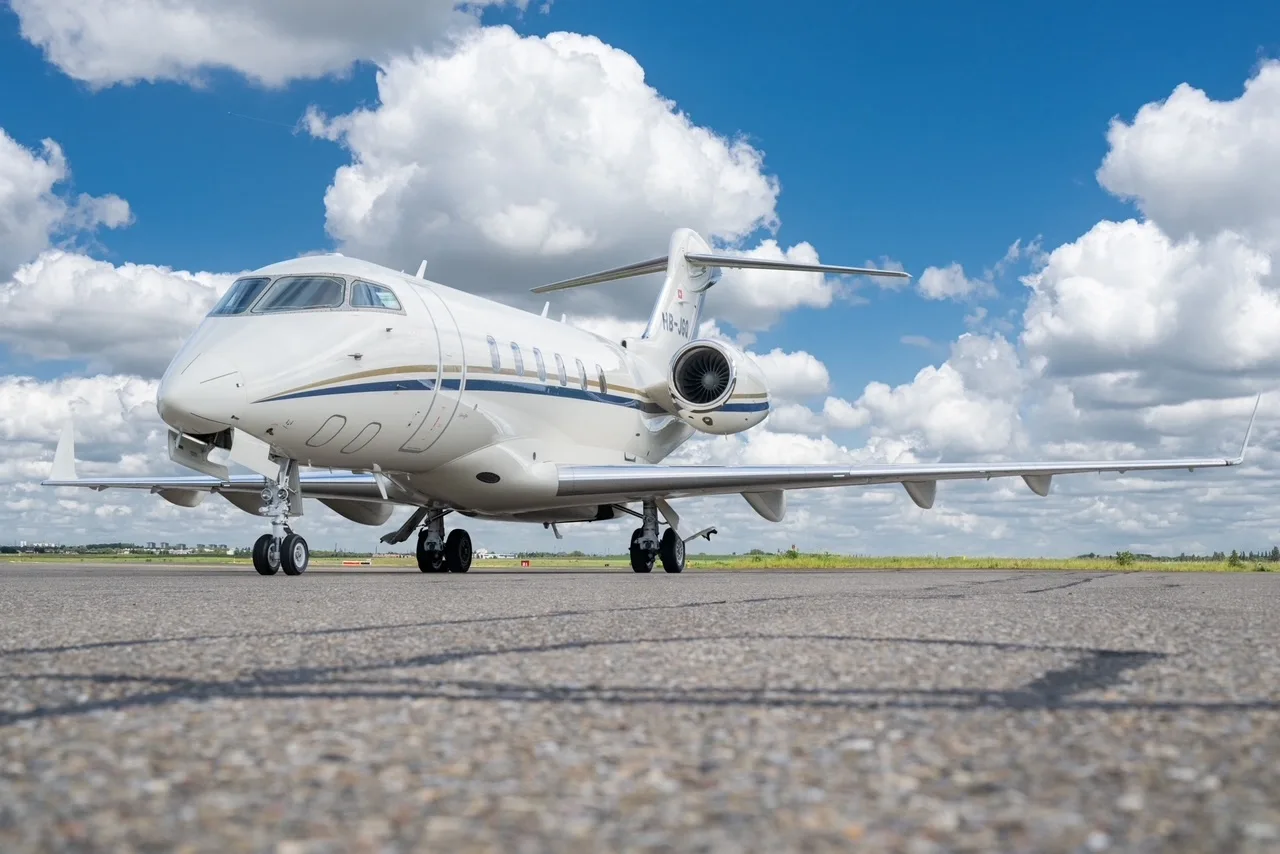A white Bombardier Challenger 300 business jet with blue and gold stripes on the tarmac under a blue sky with clouds.