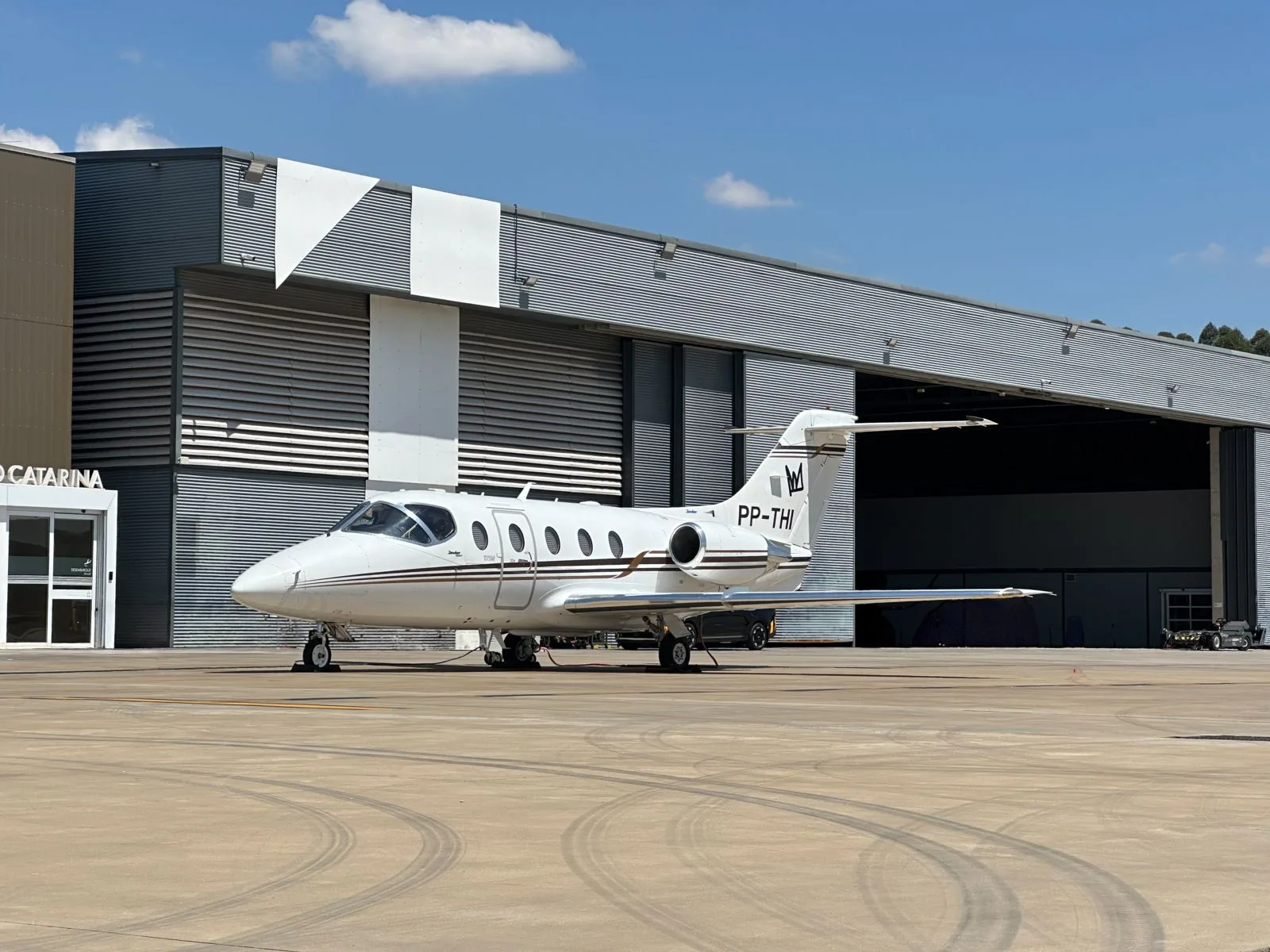 A white Learjet business jet with brown stripes is parked on an airport apron in front of a hangar under a blue sky.