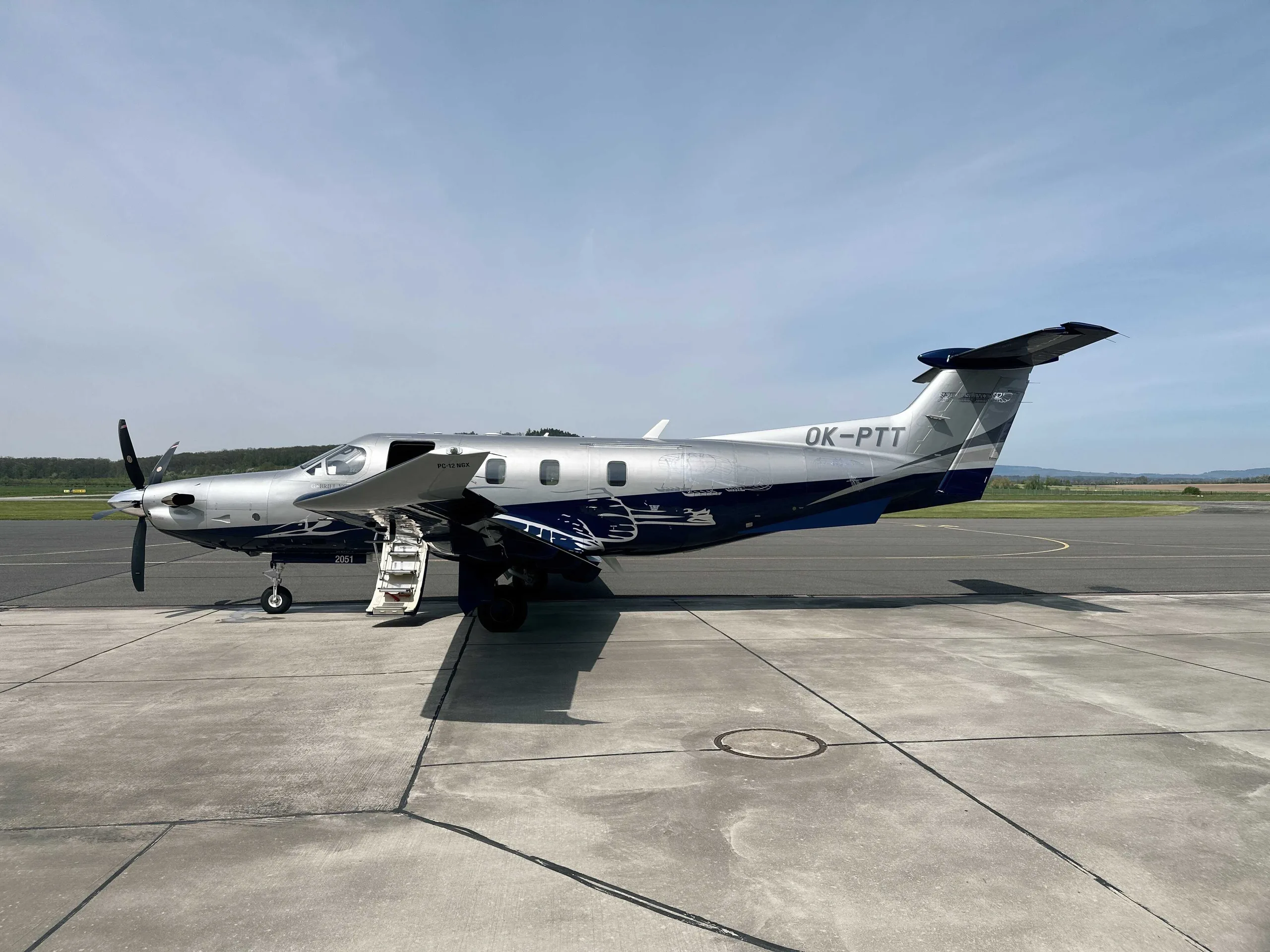 A silver and blue Pilatus PC-12 NGX turboprop aircraft with its door open, parked on an airport tarmac under a clear sky.
