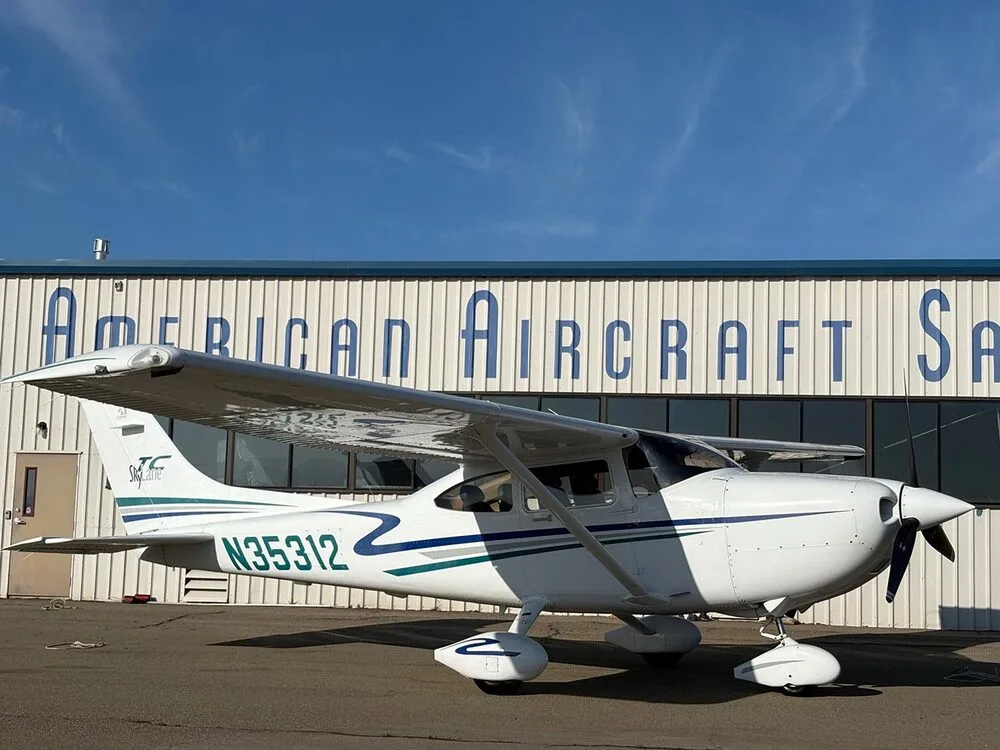 A white Cessna 182 Skylane with blue and green stripes, registration N35312, parked on an tarmac in front of a building labeled "American Aircraft Sale".