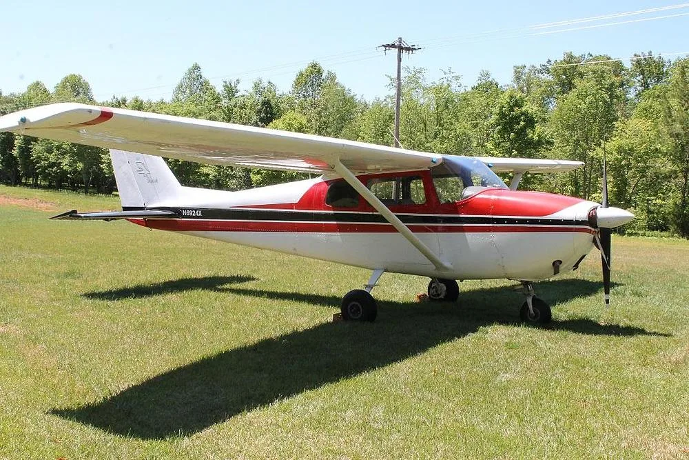 A side view of a red and white Cessna 172 (N6924X) with a black stripe, parked on a sunny grassy field.