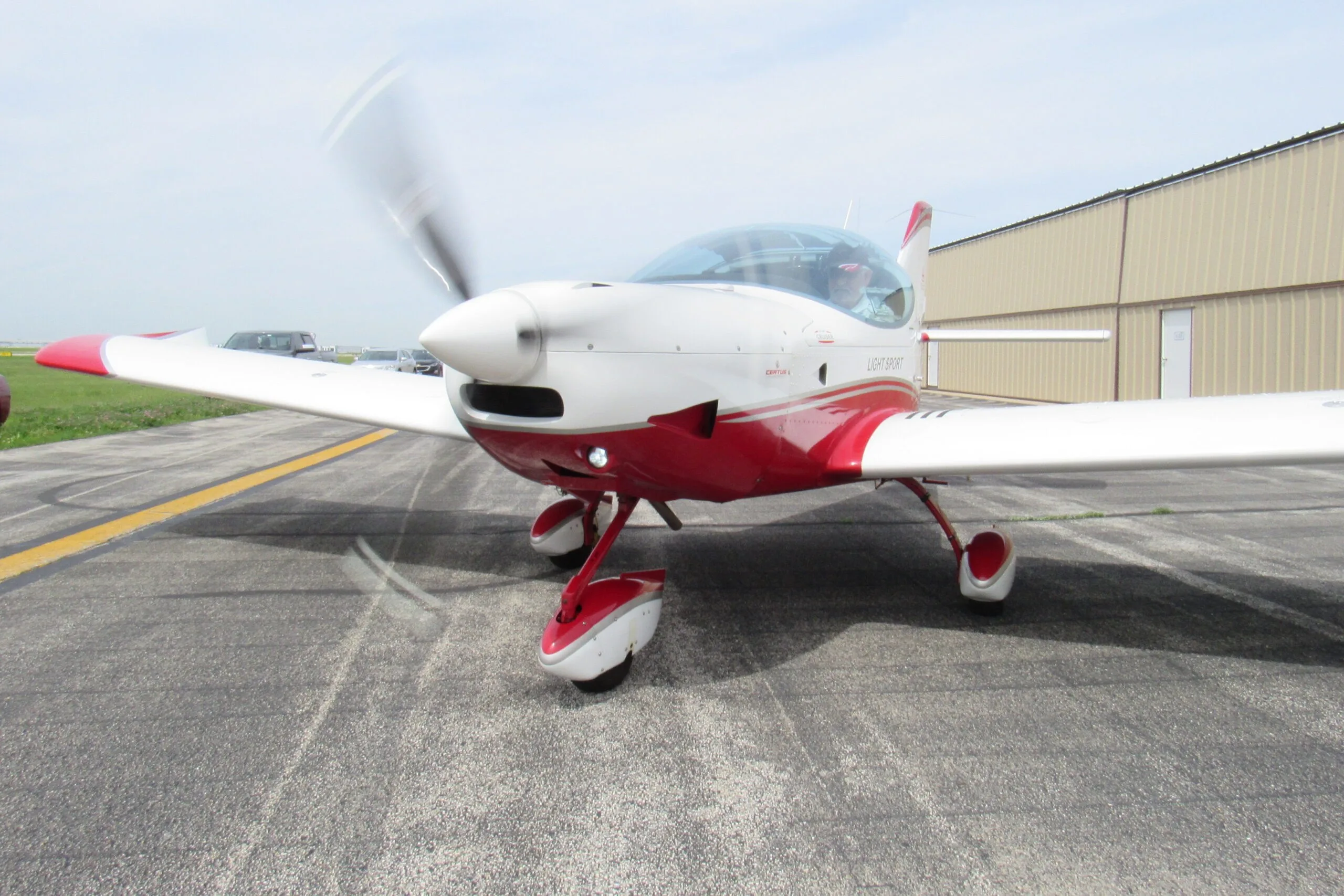 A white and red SportCruiser aircraft with a spinning propeller and a person in the cockpit on an airport tarmac.