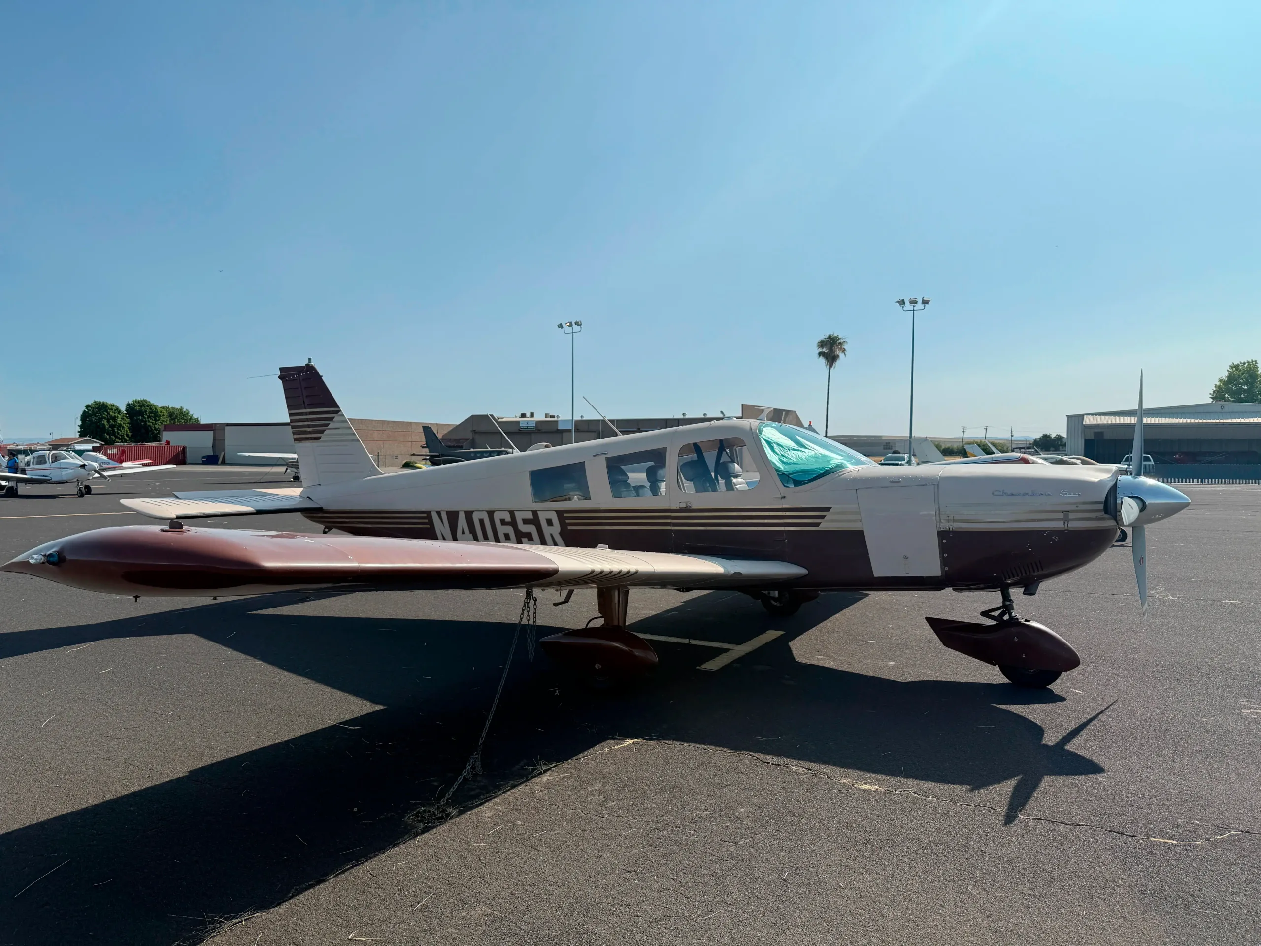 A brown and white Piper Cherokee Six aircraft, registration N4065R, is parked on an asphalt apron at an airport on a sunny day.
