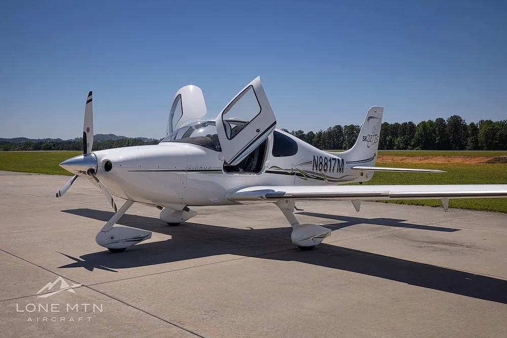 A white Cirrus SR22T aircraft (N8817M) with doors open, parked on a tarmac with trees in the background.