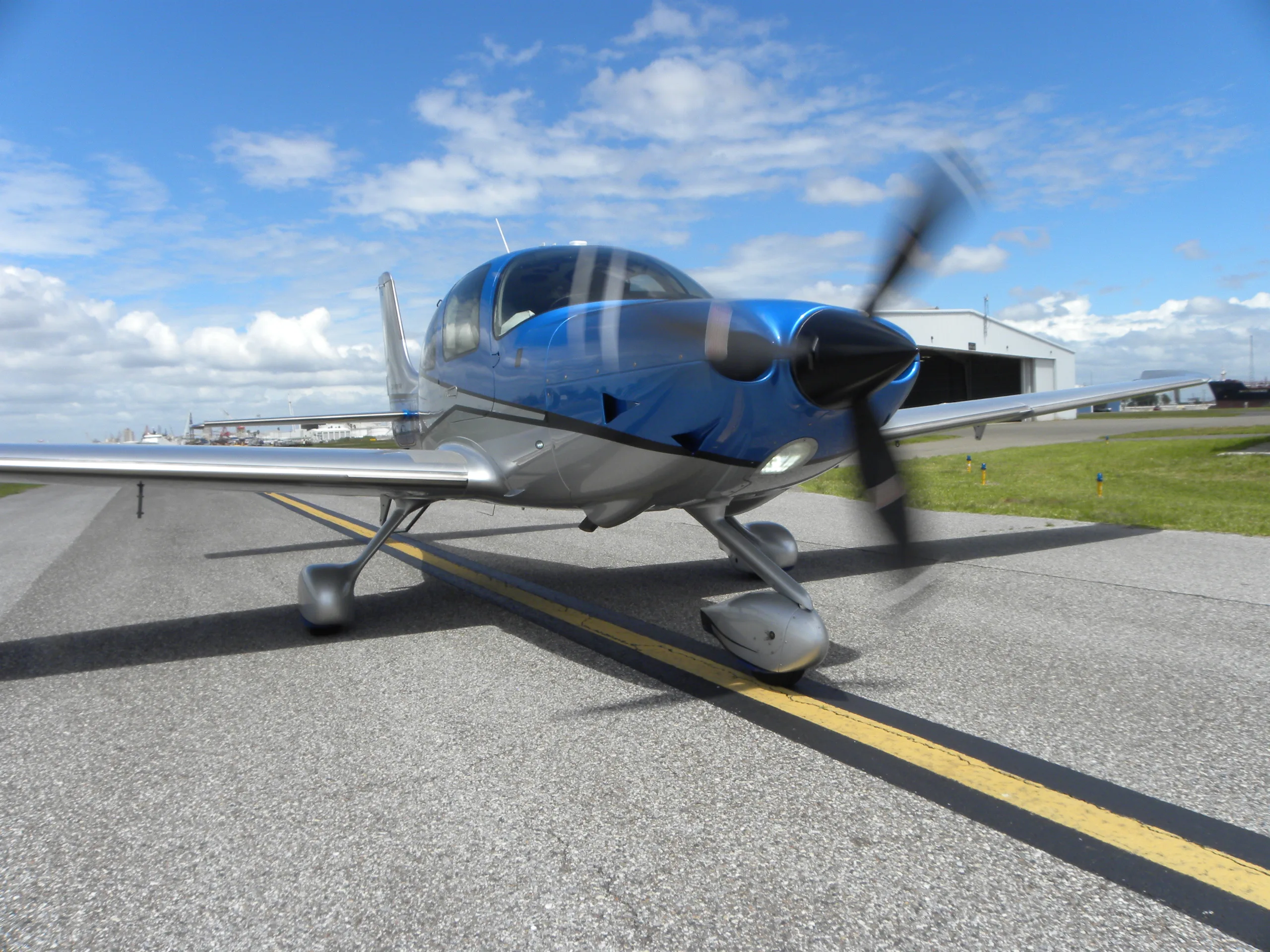 A blue and silver single-engine aircraft with a spinning propeller sits on an airport tarmac under a partly cloudy sky.