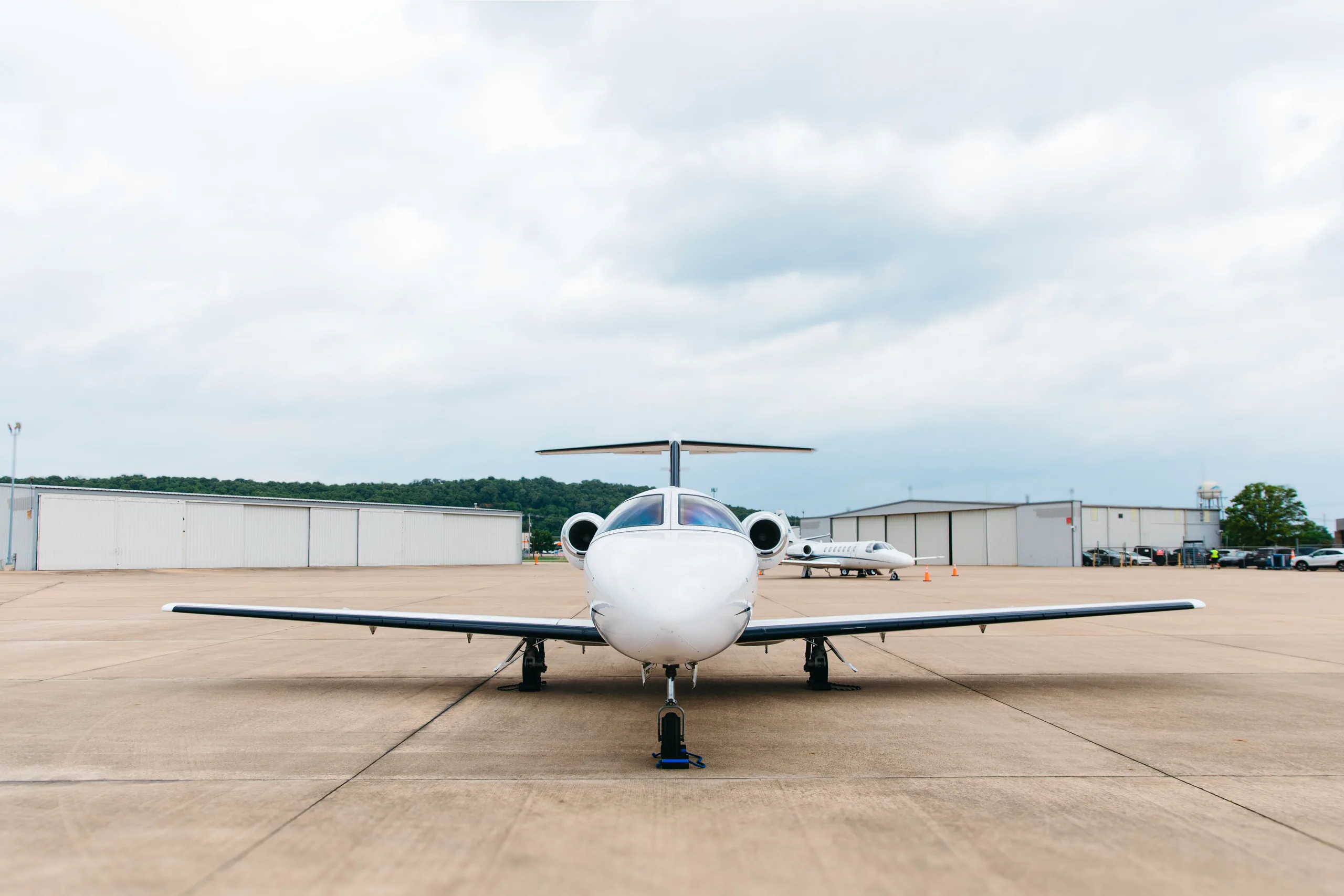 A frontal view of a white Cessna Citation M2 business jet, tail number N22EM, parked on a tarmac with another jet and hangars in the background.