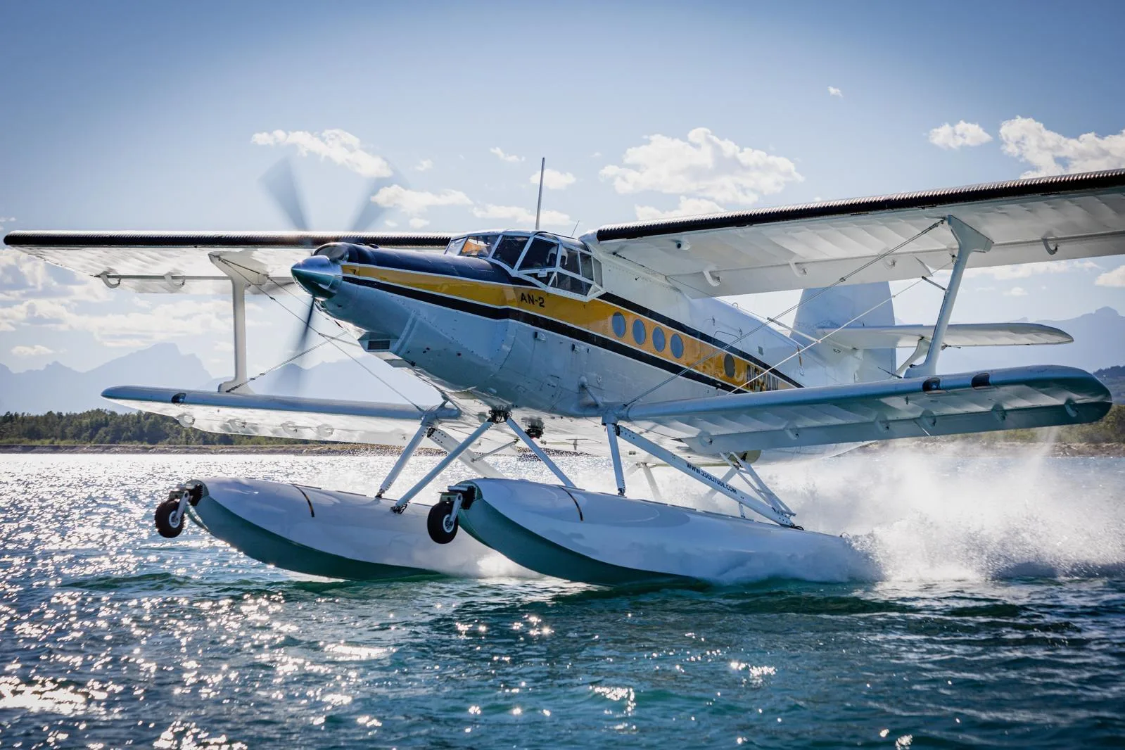 A white and yellow Antonov AN-2 floatplane takes off from a lake, creating a large splash.