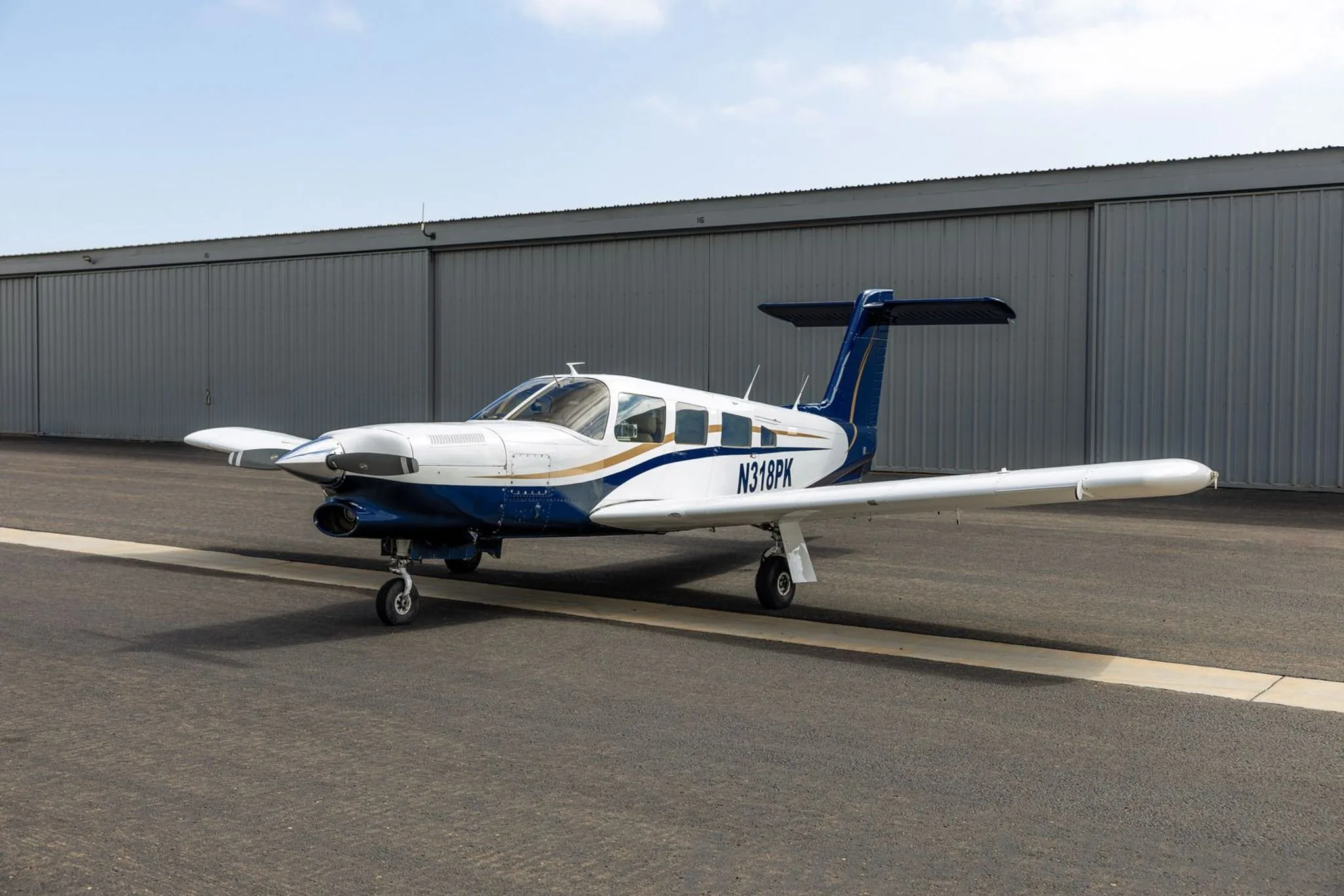 A white and blue Piper aircraft with gold accents, registration N318PK, parked on an airport tarmac in front of a hangar.