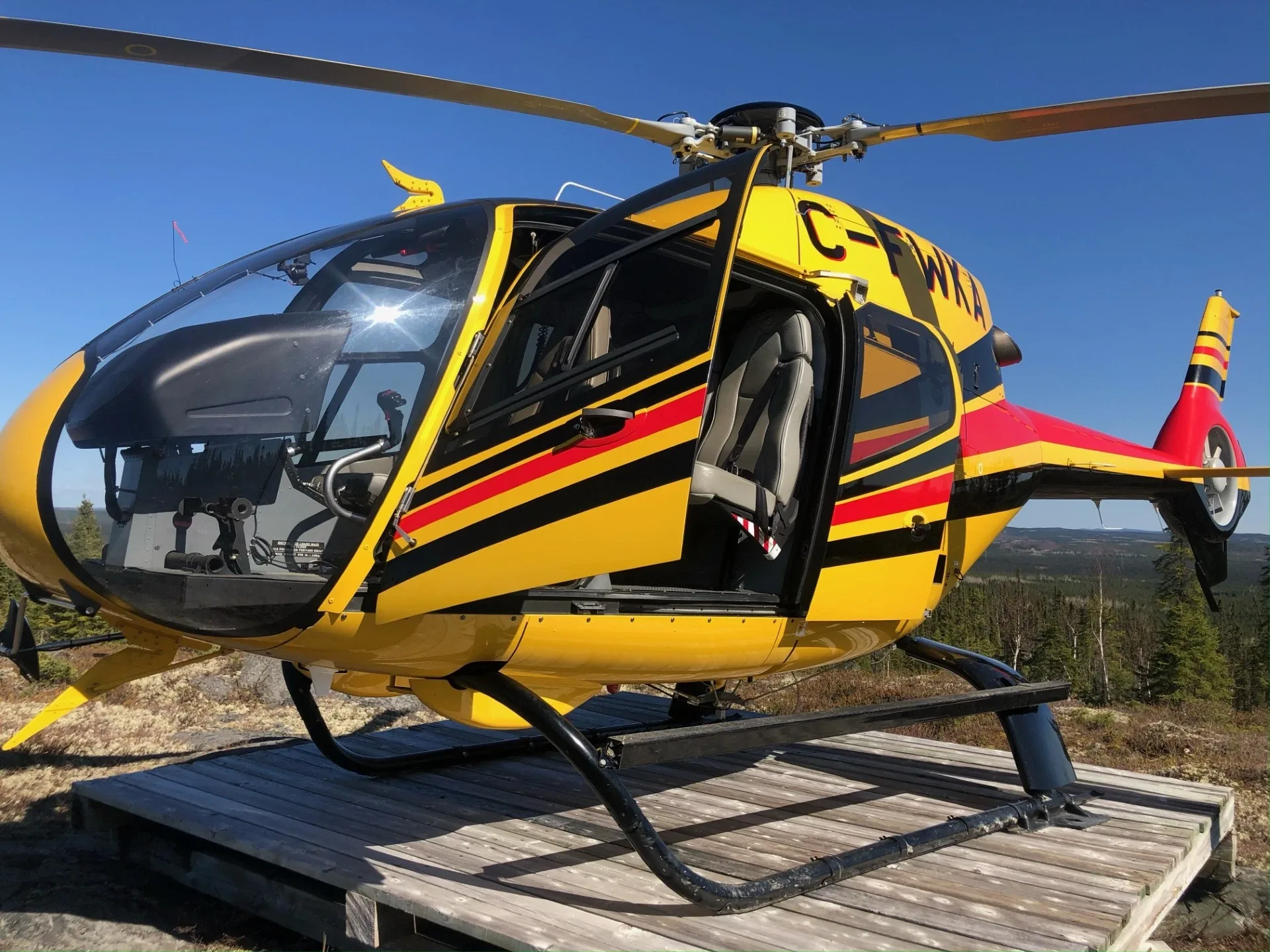 A yellow helicopter with red and black stripes, registration C-FWKA, parked on a wooden platform outdoors with its door open.