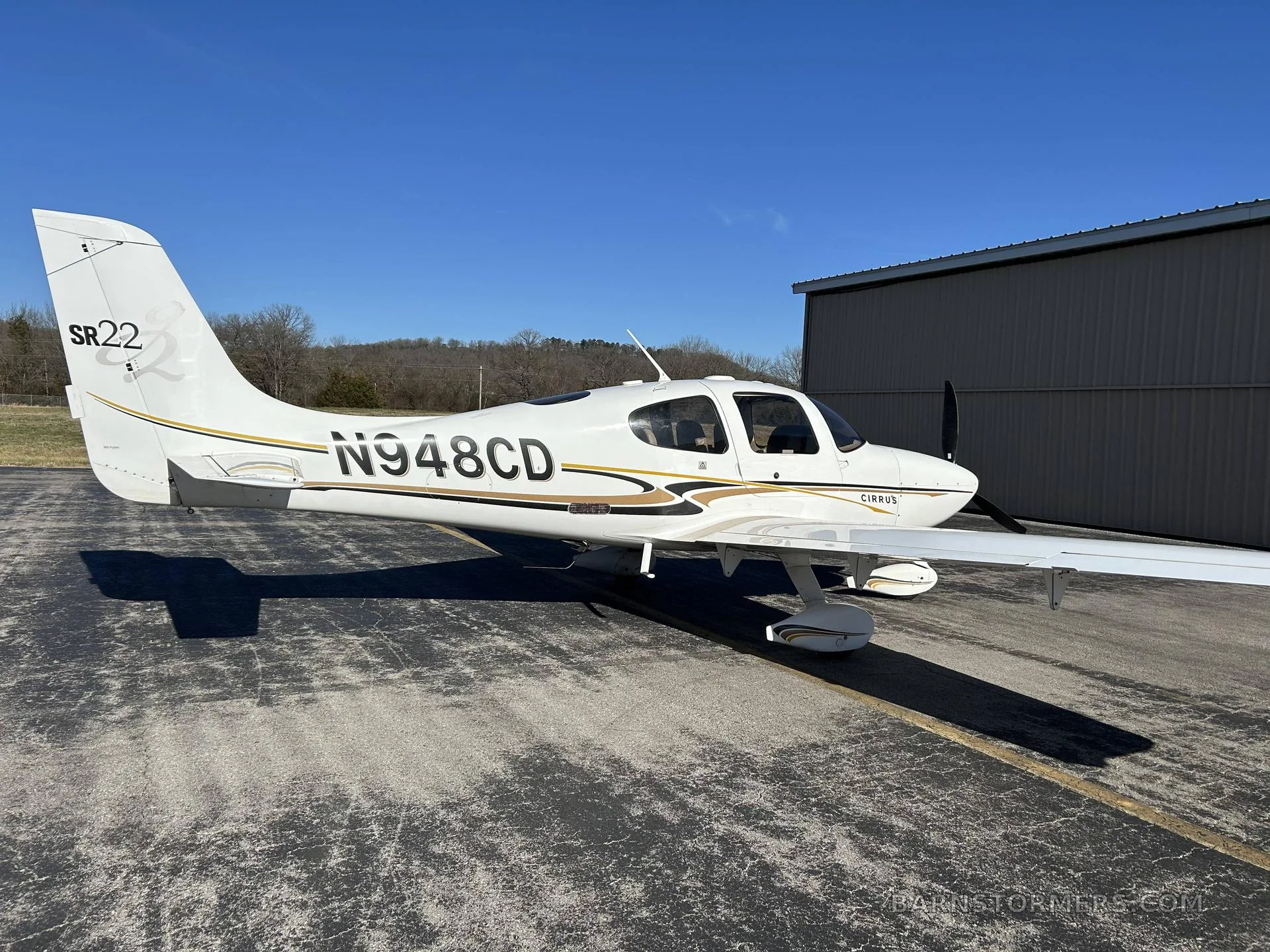 A white Cirrus SR22 aircraft with gold and black stripes, registration N948CD, parked on an asphalt ramp with a dark hangar in the background.