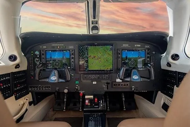 The modern glass cockpit of a small aircraft, featuring multiple display screens and controls, with a sunset sky visible through the windshield.