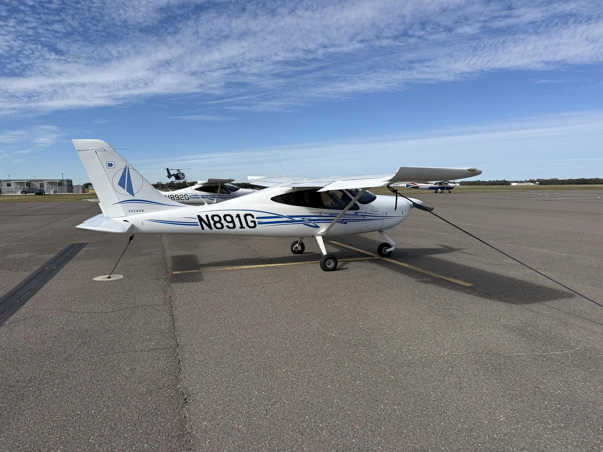 A white Tecnam P2008 aircraft with blue stripes and N891G registration, viewed from the front-right on an airport tarmac.