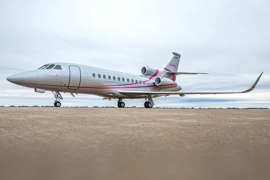 A low-angle side view of a silver Dassault Falcon 900EX private jet with pink accents on a concrete surface.