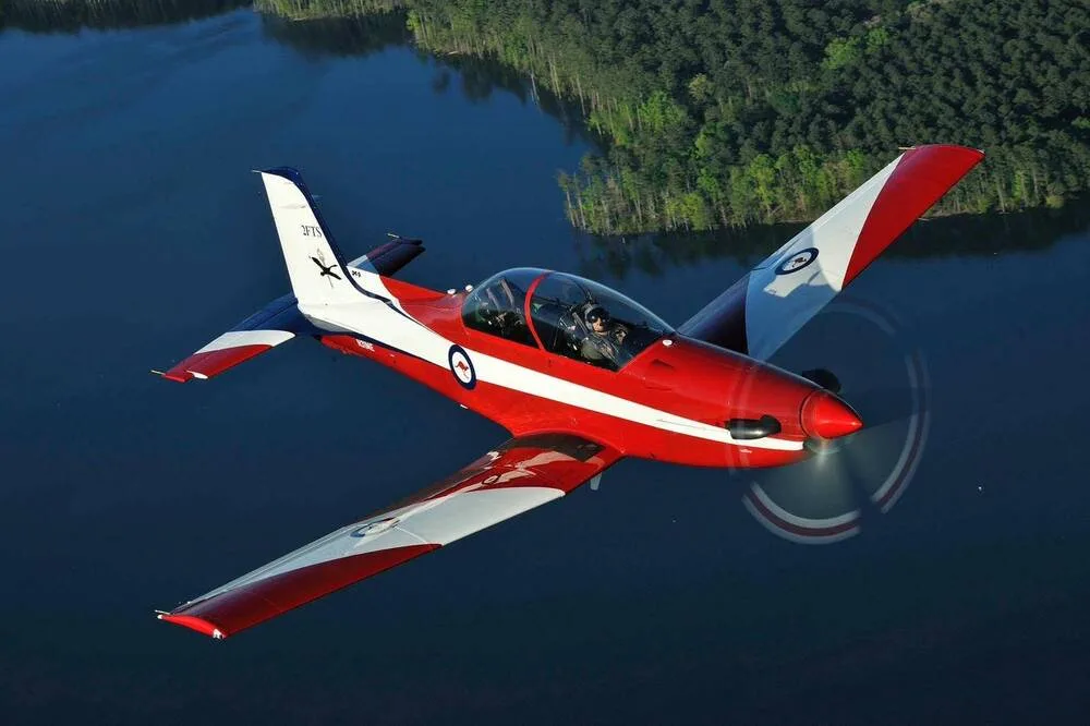A red and white single-engine aircraft with a bubble canopy flies over a lake and forest.