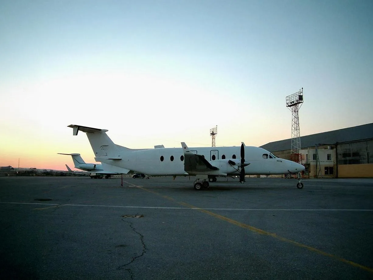 A white twin-engine turboprop aircraft is parked on an airport tarmac at sunset, with another aircraft in the background.