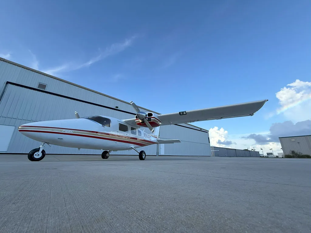 A white and red twin-engine aircraft, N564BC, is parked on an airport tarmac with hangars and a faint rainbow in the sky.