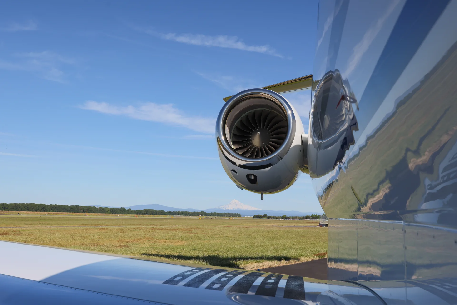 A close-up of the polished engine and wing of a white business jet, reflecting the blue sky, with a distant mountain.
