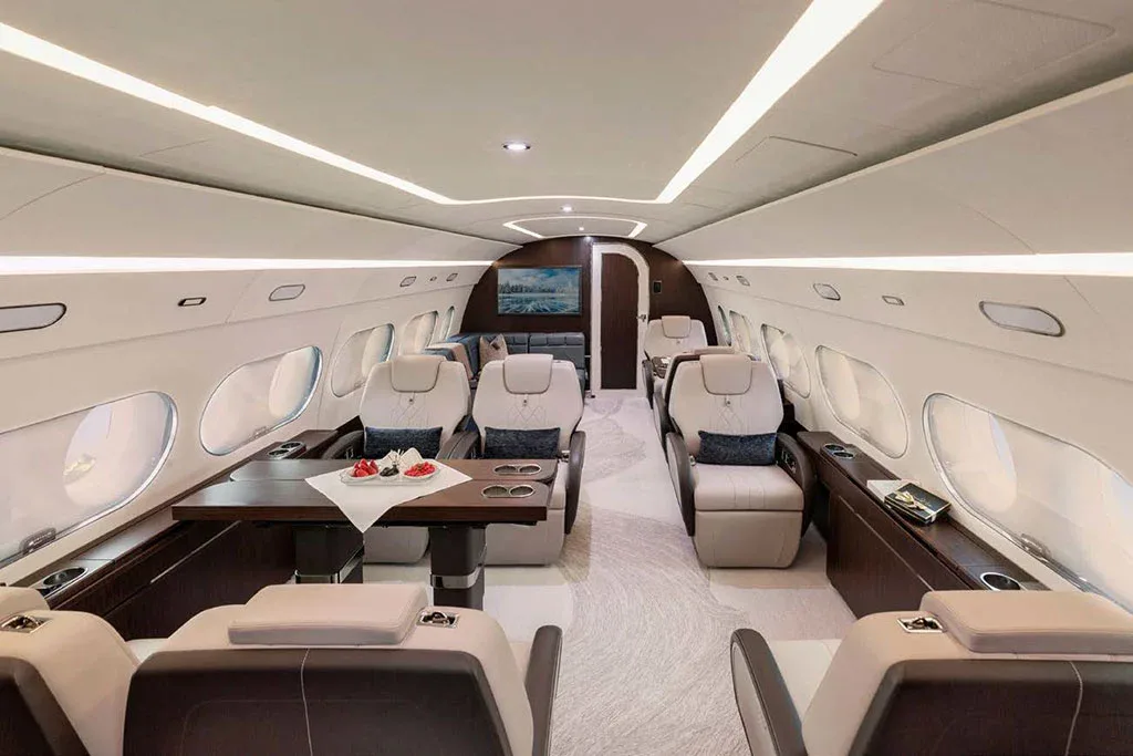 A wide view of a private jet cabin with light beige seats, a central dining table, and a TV screen at the far end.