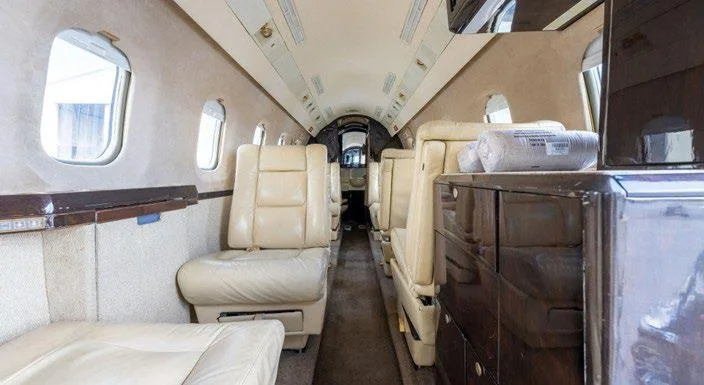 The interior cabin of a business jet with beige leather seating and dark wood cabinetry.