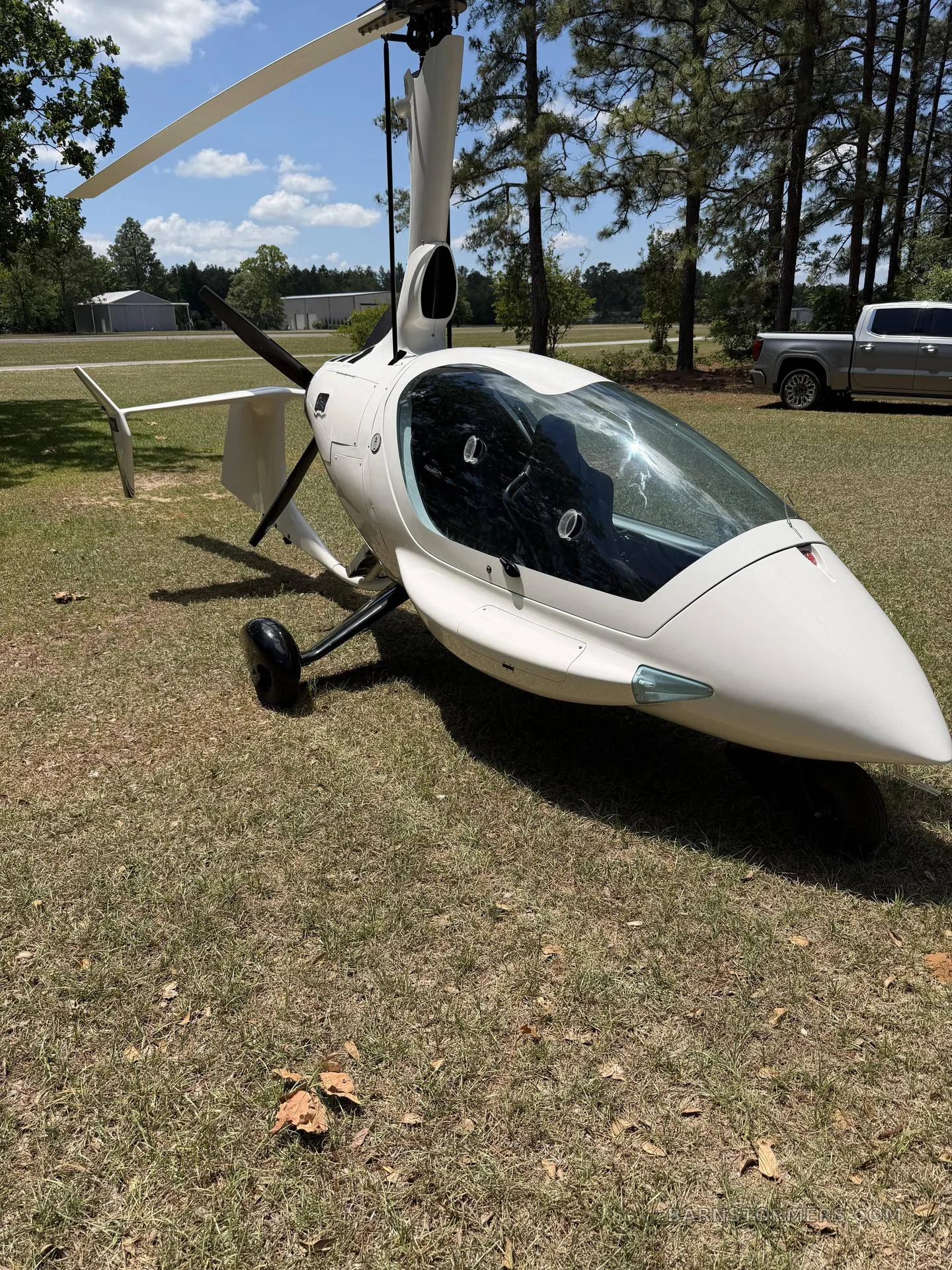 A white two-seat autogyro with a large clear canopy is parked on a grassy field with trees and buildings in the background.